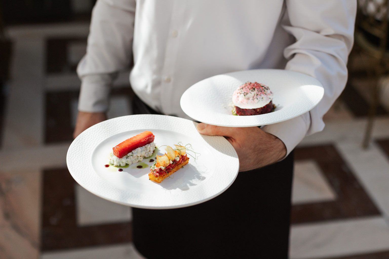 Číšník Villa Richter Restaurant nese dva předkrmy z degustačního menu – pstruha s hash brown, okurkou, koprem a kaviárem a tatarák z červené řepy s crème fraîche