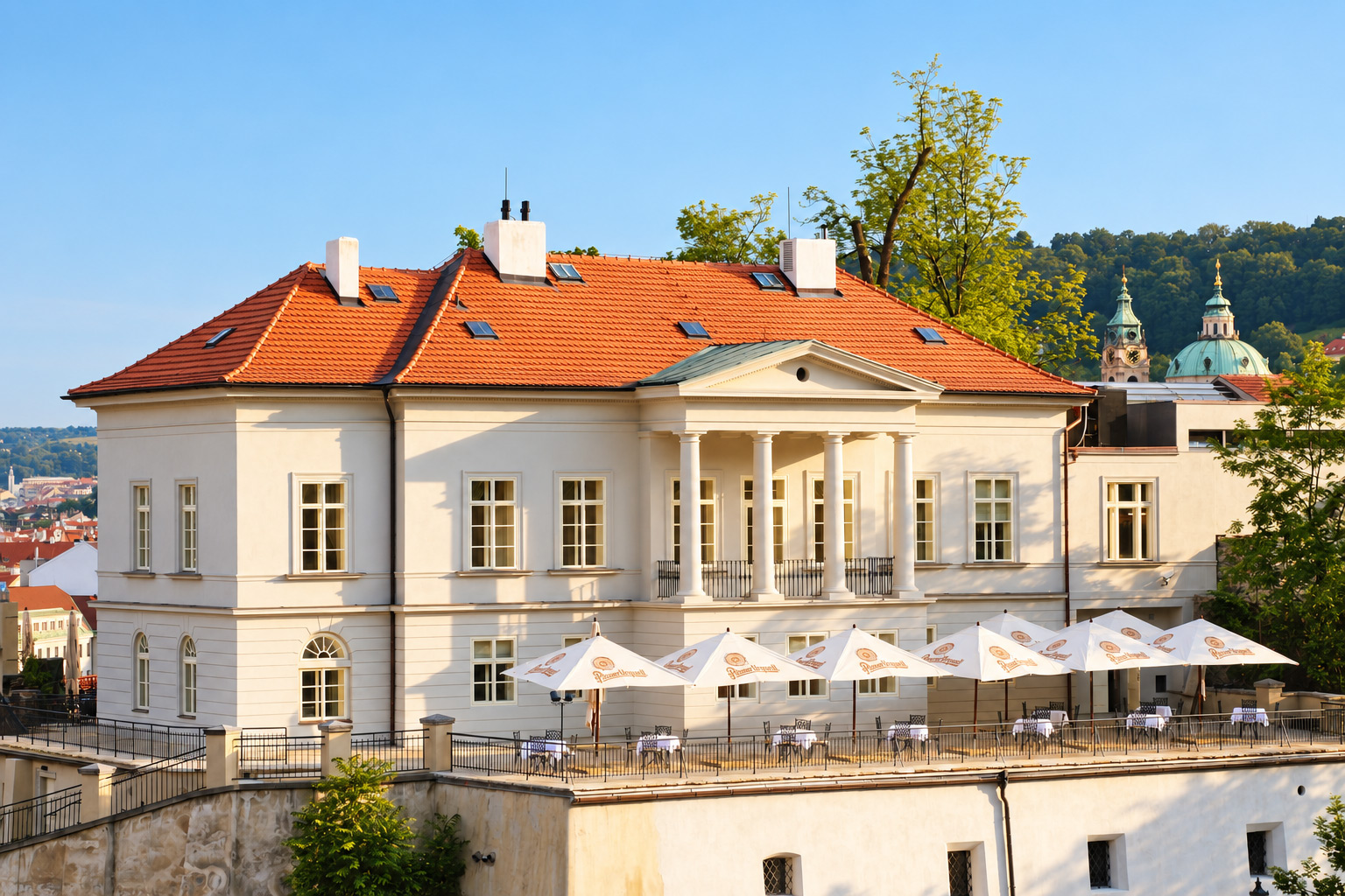 Klasicistní budova Villa Richter s terasou pod slunečníky, v pozadí kopule kostela sv. Mikuláše na Malé Straně. Pohled z Chotkových sadů